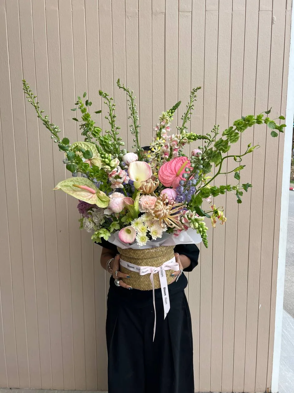 Lush pastel arrangement in a woven seagrass basket with pink and green anthuriums, pink peonies, purple delphinium, snapdragons, bells of Ireland, white chrysanthemums and a calla lily