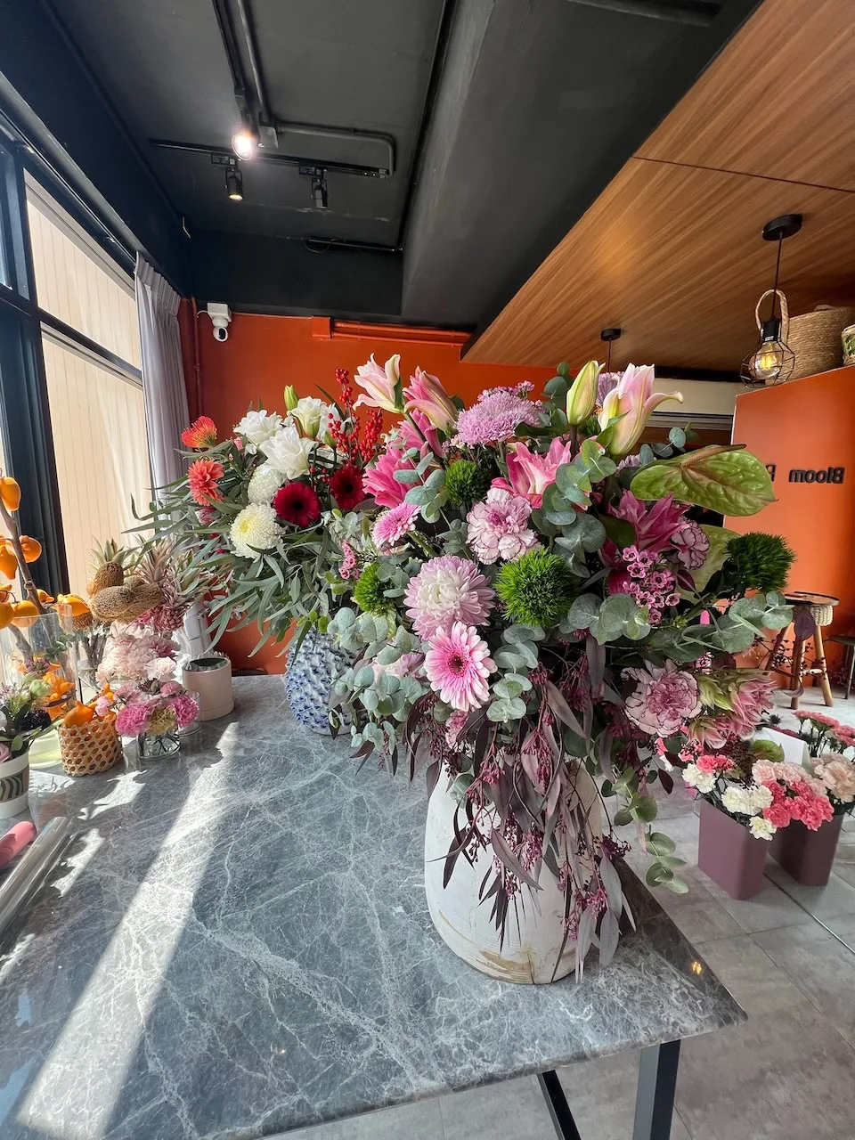 Oversize arrangement of pink lilies, pink gerberas, green chrysanthemums, lavender mums, eucalyptus, and trailing amaranth in a white ceramic vase on the studio counter.