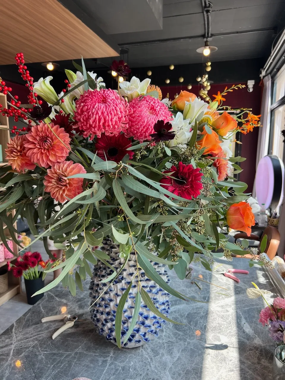Large studio arrangement of hot pink chrysanthemums, coral dahlias, red ilex berries, white lilies, orange crocosmia, and cascading eucalyptus in a blue-and-white ceramic vase.