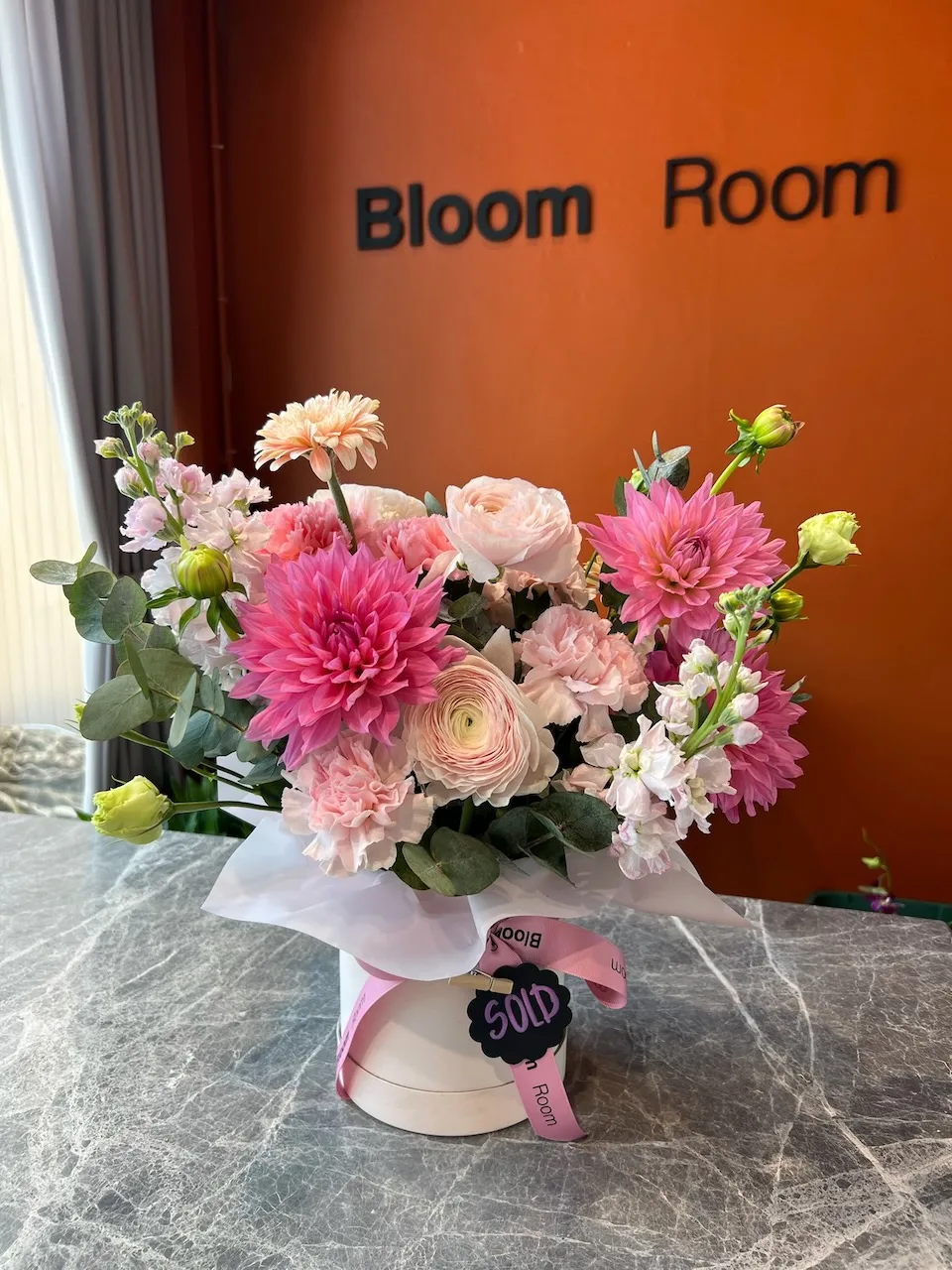 Pink dahlia, ranunculus and chrysanthemum hatbox with Bud tag on marble under Bloom Room sign
