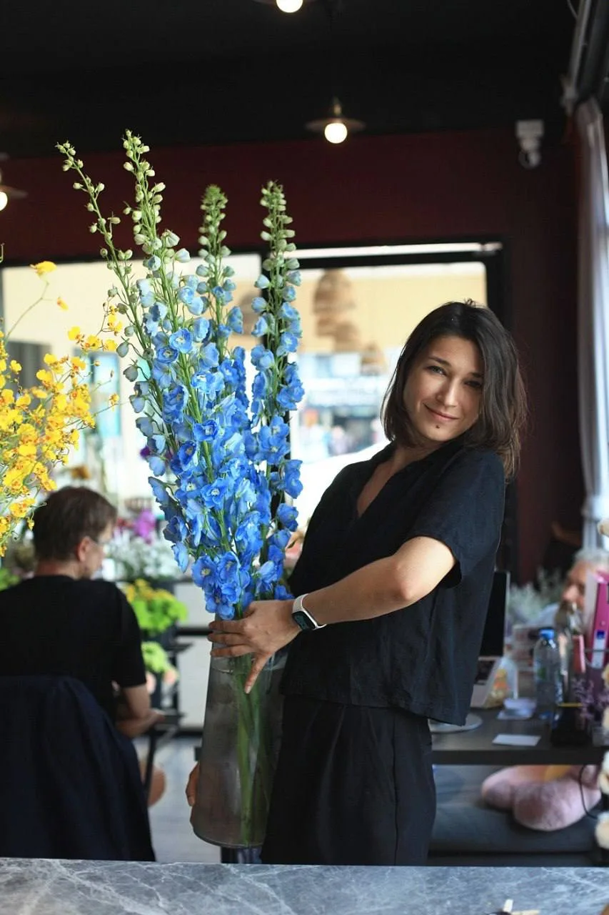 A florist in black holds a tall glass vase of bright blue delphinium at the studio counter, with yellow oncidium orchids nearby.
