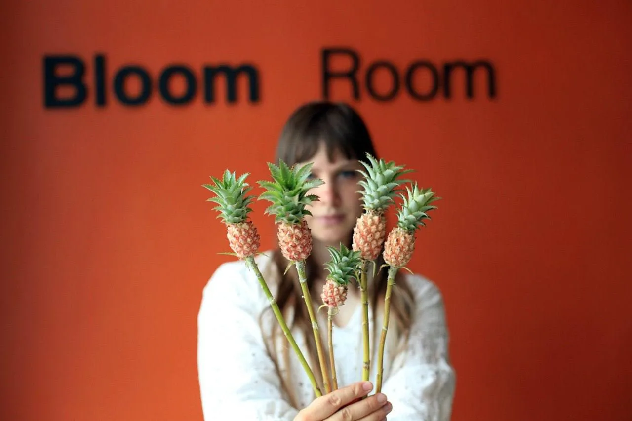A woman holds a bunch of ornamental pineapples in front of the orange Bloom Room sign.